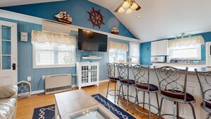 A nautical-themed living room with a large TV, bar stools, and a ship model on the wall. - Sand Life #2 by Bayley Vacation Rentals (Old Orchard Beach)