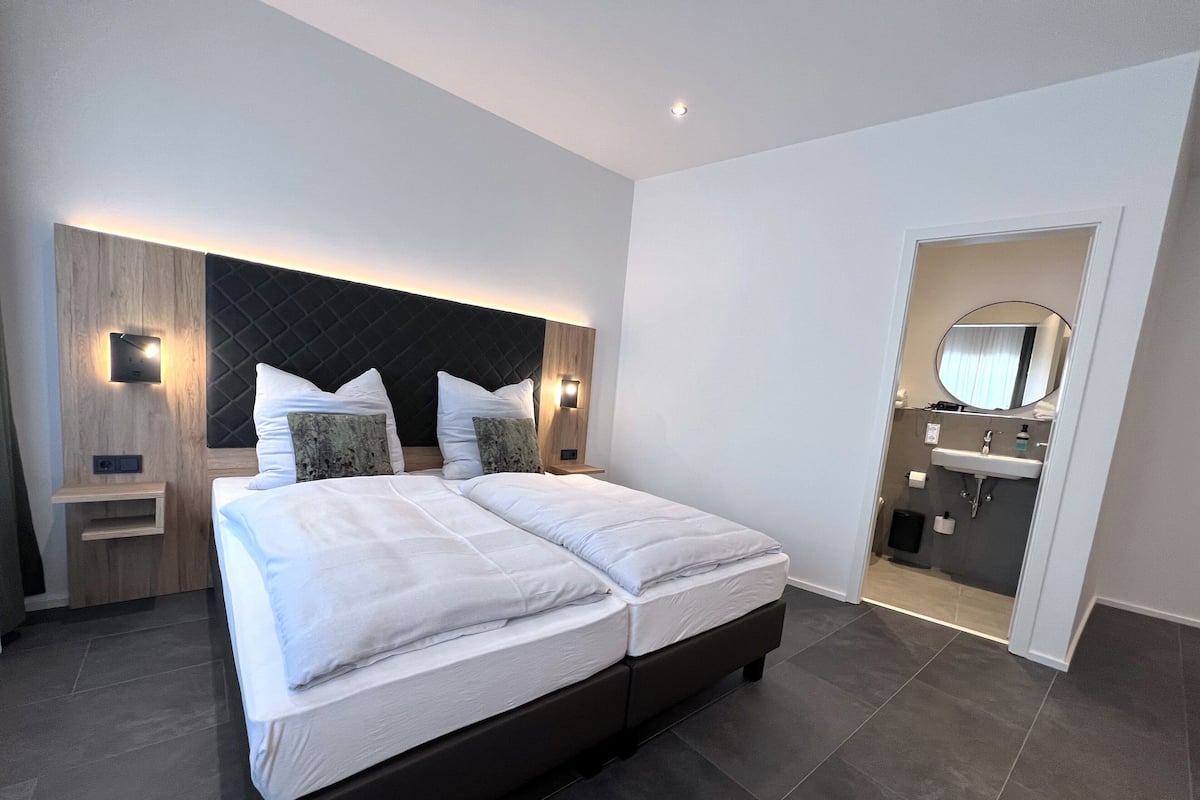 A modern hotel room with a large bed, a built-in nightstand, a wall-mounted light, and a bathroom visible through an open door.