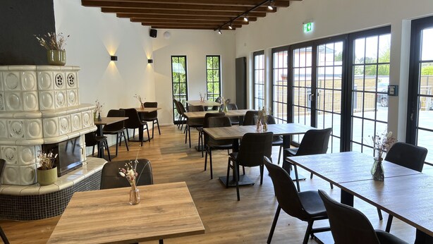 A modern dining area with wooden tables and chairs, a decorative fireplace, and large windows offering views of the outdoors. - Haus Kuckenberg (Burscheid)