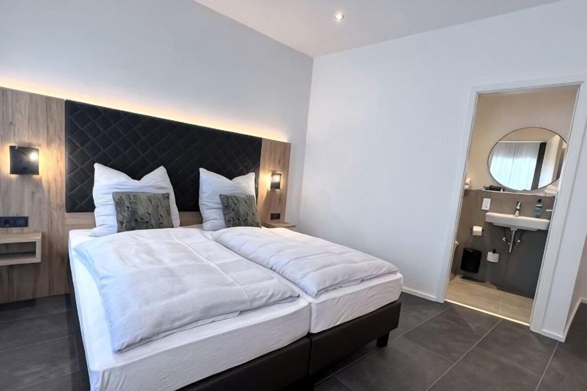 A modern hotel room with a large bed, bedside tables, a built-in headboard, a bathroom with a mirror and sink.