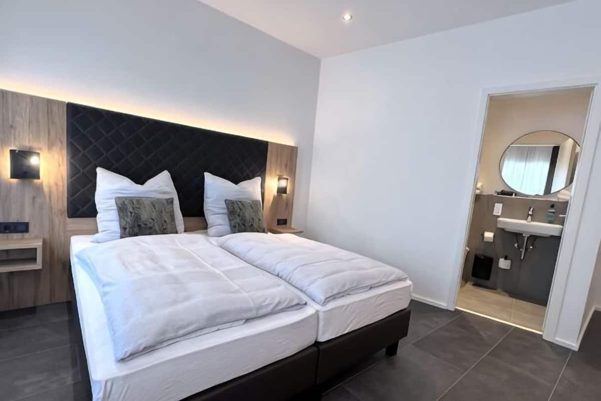 A modern hotel room with a large bed, bedside tables, a built-in headboard, a bathroom with a mirror and sink.