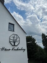 Um edifício com fachada branca, um logotipo hexagonal com desenho de árvore e o texto "Haus Kuckenberg" na lateral.