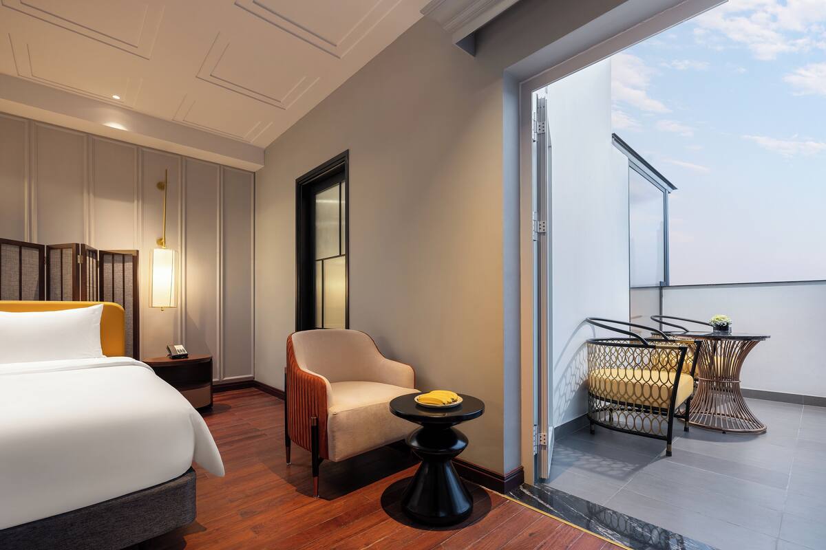 A hotel room with a bed, a chair, a small table, and a balcony with a table and chairs.