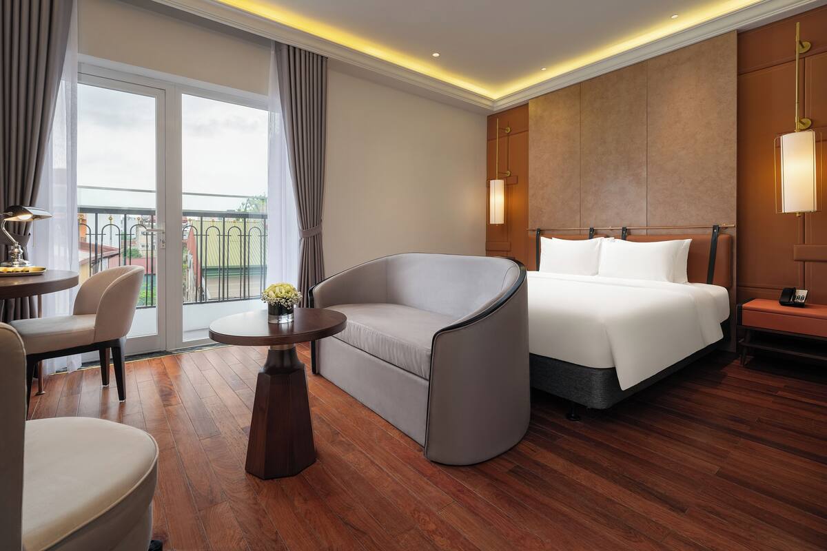 A hotel room with a bed, a sofa, a small table with a lamp, and a view of the outdoors through a large window.