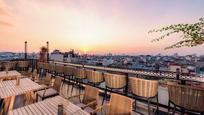 Eine Dachterrasse mit Korbmöbeln und Tischen, die einen Blick auf die Stadt bei Sonnenuntergang bietet.
