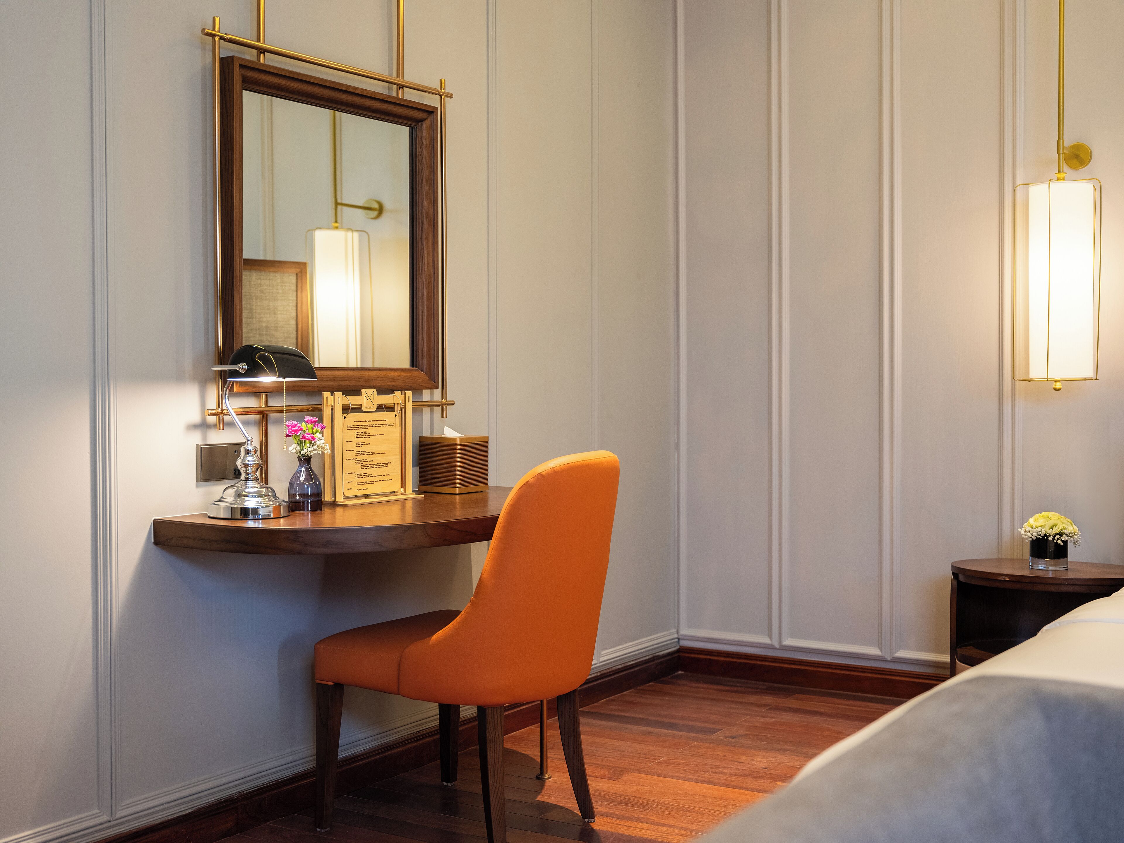 A hotel room with a wooden desk, a chair, a bed, a nightstand, a lamp, and a wall-mounted mirror.