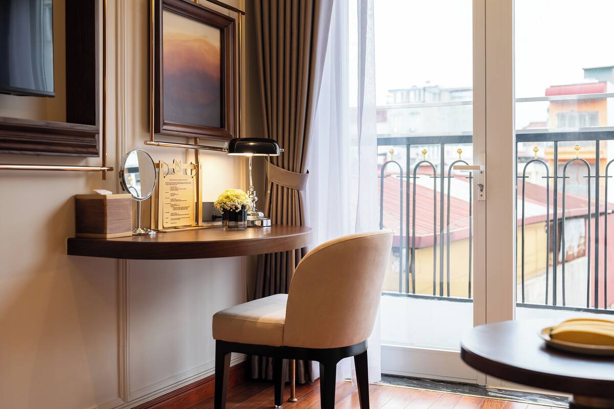 A modern hotel room with a wooden desk, a chair, a bedside table with a lamp, a framed picture, and a view of the city through a balcony with a metal railing.