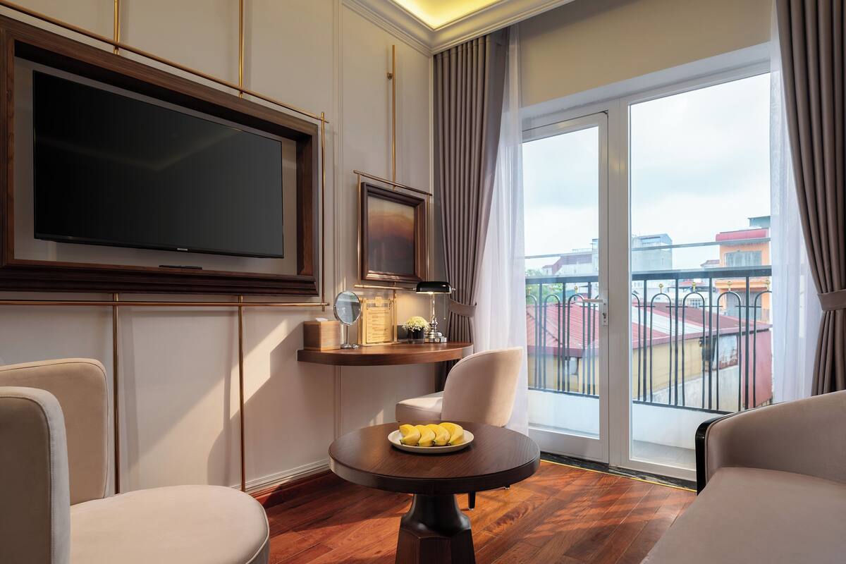 A hotel room with a large flat-screen TV, a round wooden table with a plate of fruit, two armchairs, and a view of the city through a balcony with a railing.