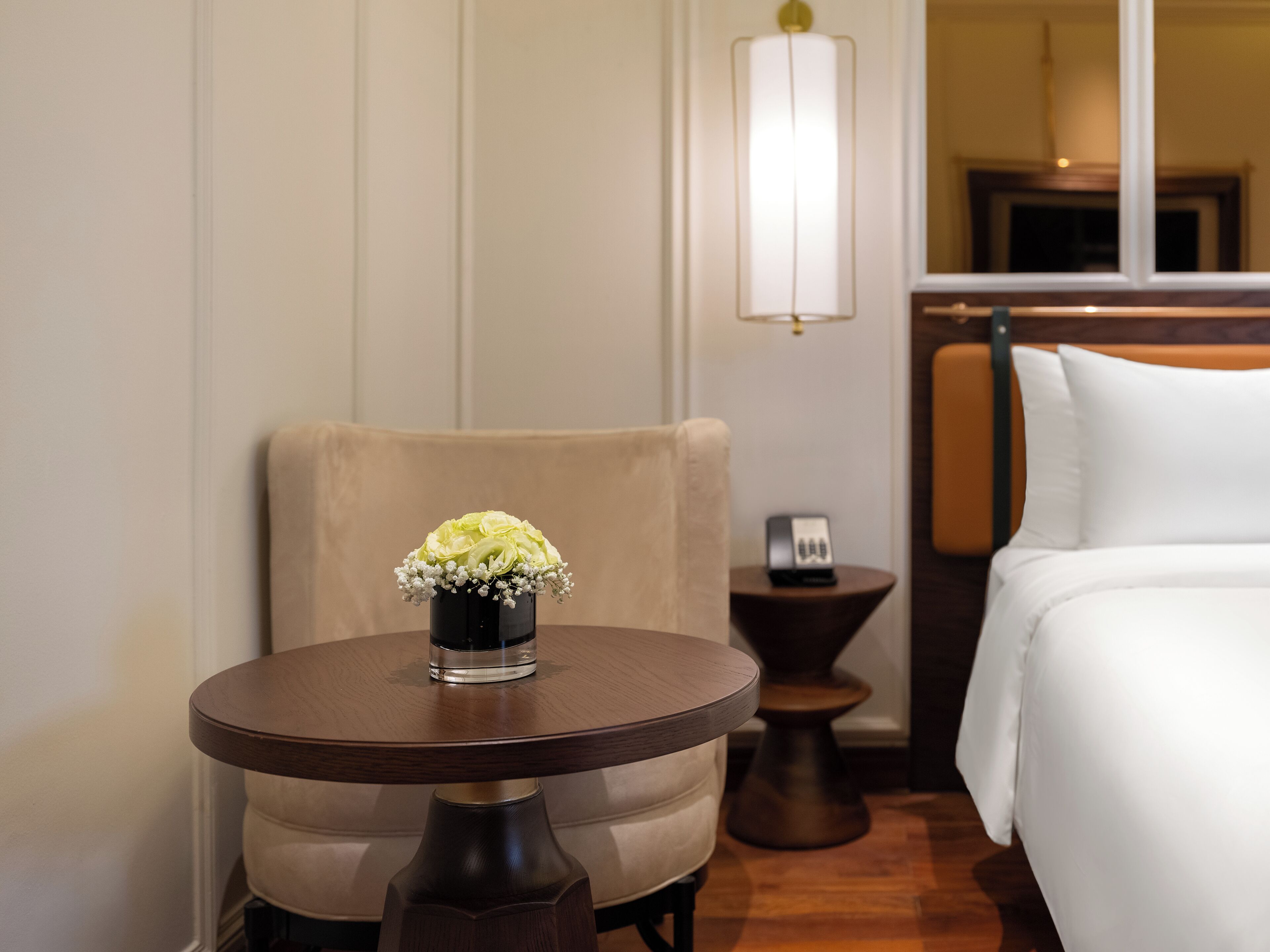 A hotel room with a bed, a round table with a vase of flowers, a chair, a bedside table, and a lamp.