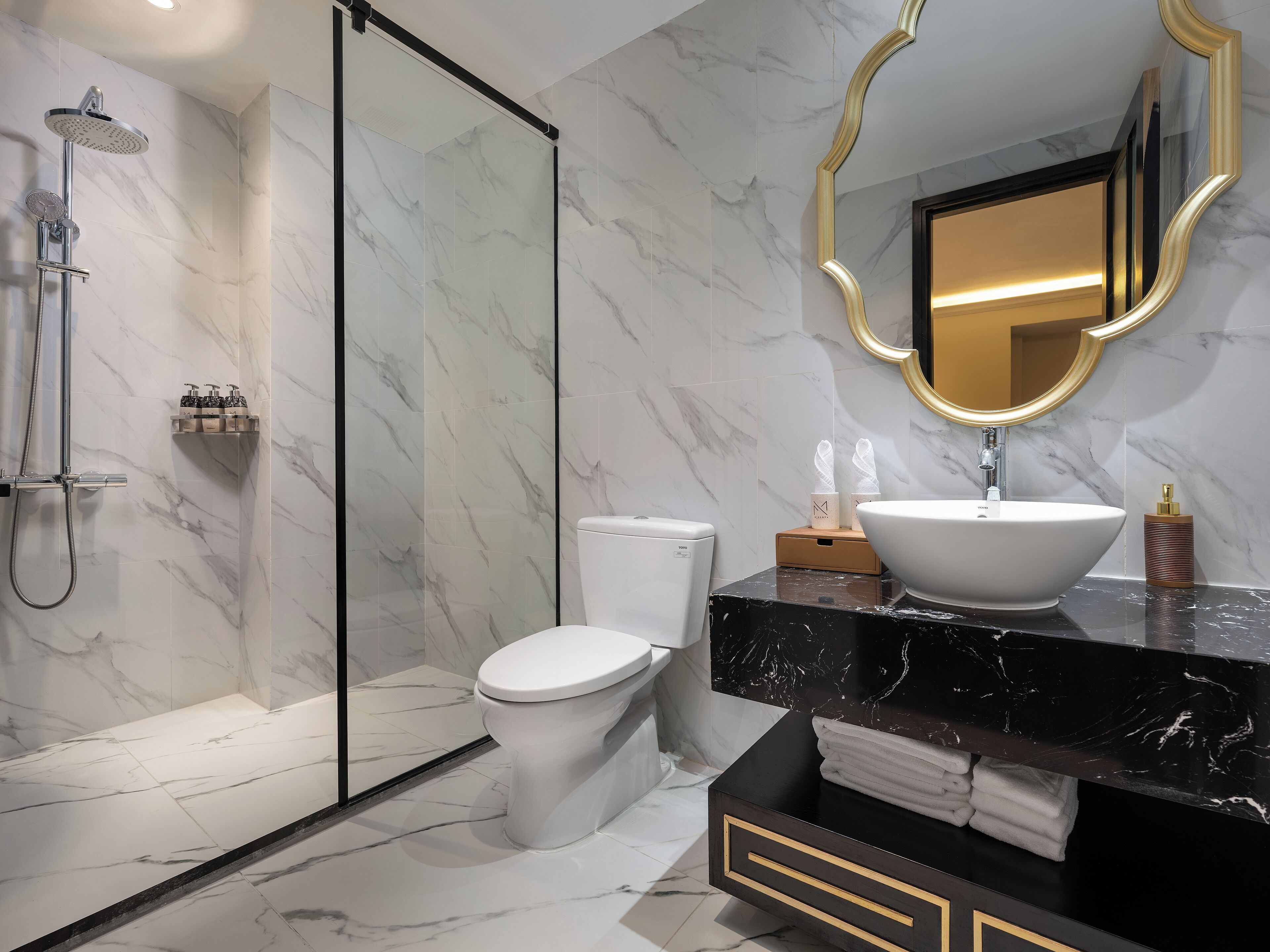 Une salle de bain moderne dotée d’une douche à parois de verre, de toilettes, d’un meuble-lavabo avec un grand évier et d’un miroir encadré d’or.