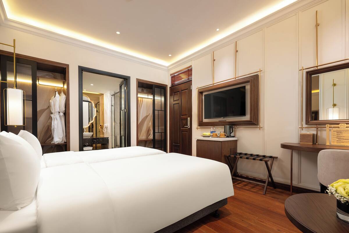 A spacious hotel room with a large bed, a desk with a flat-screen TV, a vanity with a mirror, and a seating area with a chair and a small table.