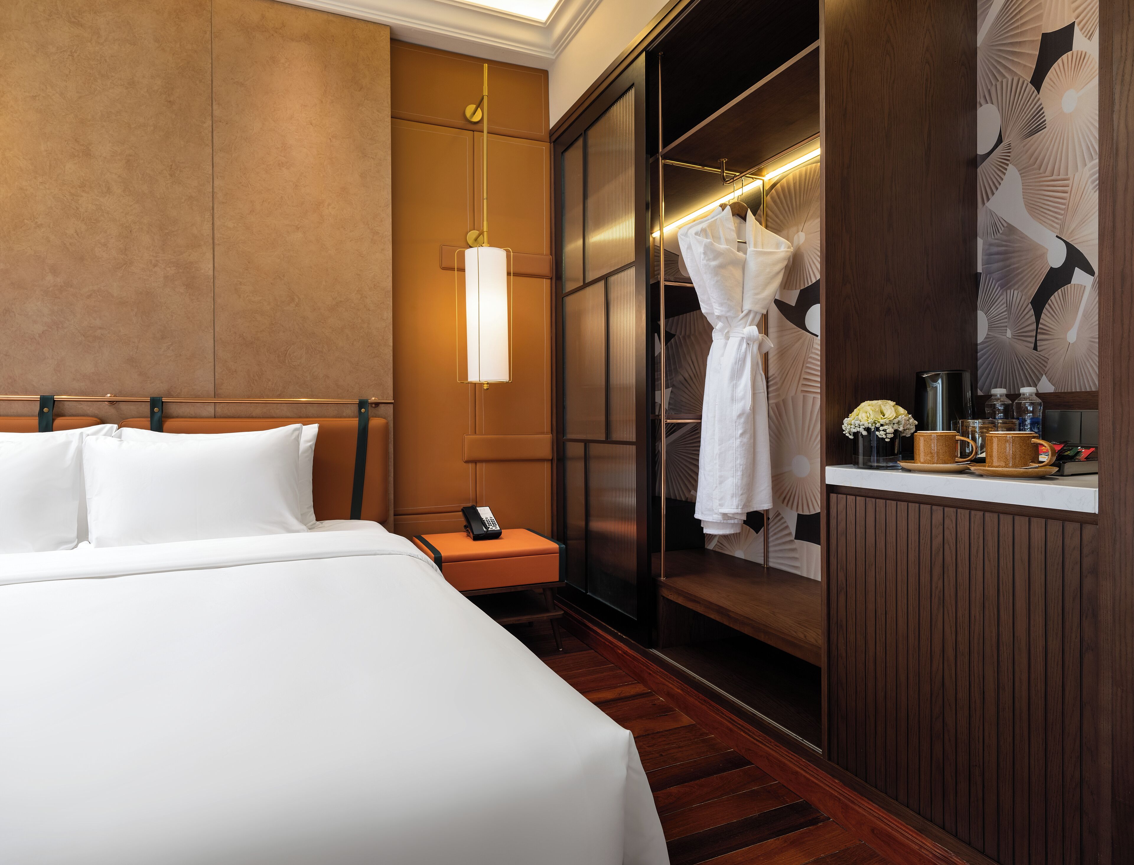 A hotel room with a large bed, a bedside table with a phone, a robe hanging in a closet, and a small table with a vase of flowers and cups.