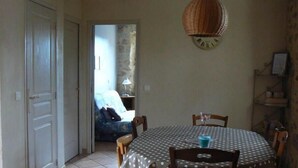 A dining area with a polka dot tablecloth, wooden chairs, a clock on the wall, and a doorway leading to a living room with a sofa and lamp.