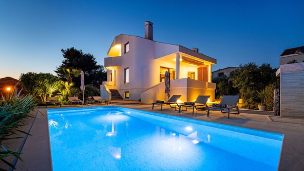 Una casa moderna de dos pisos con piscina, sillas de descanso y un espacio exterior bien iluminado.