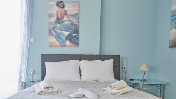 Una cama bien hecha con ropa blanca y toallas, una mesita de cristal con una lámpara, y un cuadro de una mujer en la pared.