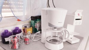 Una cafetera con depósito de agua, una caja de filtros de papel y frascos de azúcar y otros condimentos sobre una encimera.