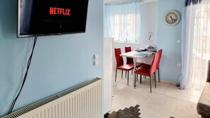 Una habitación con una televisión que muestra 'NETFLIX', un comedor con sillas rojas, una mesa blanca y un reloj en la pared.