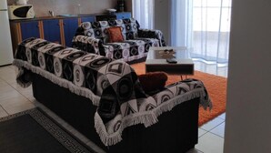A living room with a sofa and a coffee table, a kitchen with blue cabinets, and a large window with curtains. - Daphnus Apartment in Agios Konstantinos (Kamena Vourla)