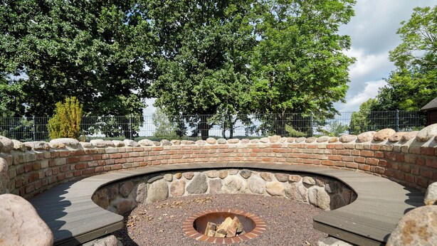 A circular stone fire pit with a brick surround, surrounded by a wooden deck and large rocks. - Ferienhaus Nationalparkidylle in Boek (Rechlin)