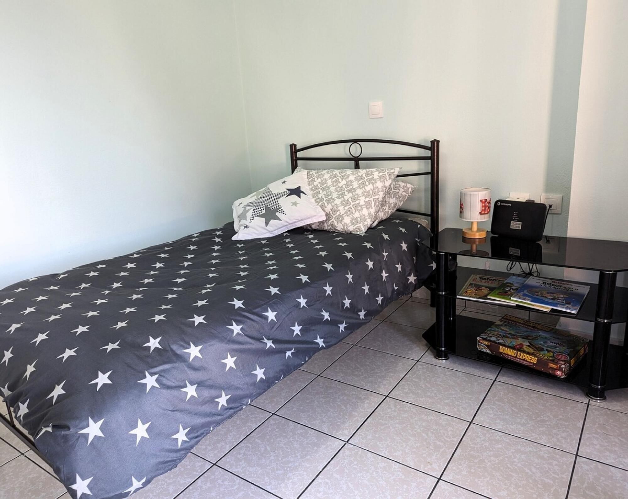 Un lit simple avec une couvre-lit à motifs d’étoiles, une table de chevet avec une lampe et une boîte de jeu Domino Express, et une prise de courant au mur.