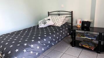 Un lit simple avec une couvre-lit à motifs d’étoiles, une table de chevet avec une lampe et une boîte de jeu Domino Express, et une prise de courant au mur.