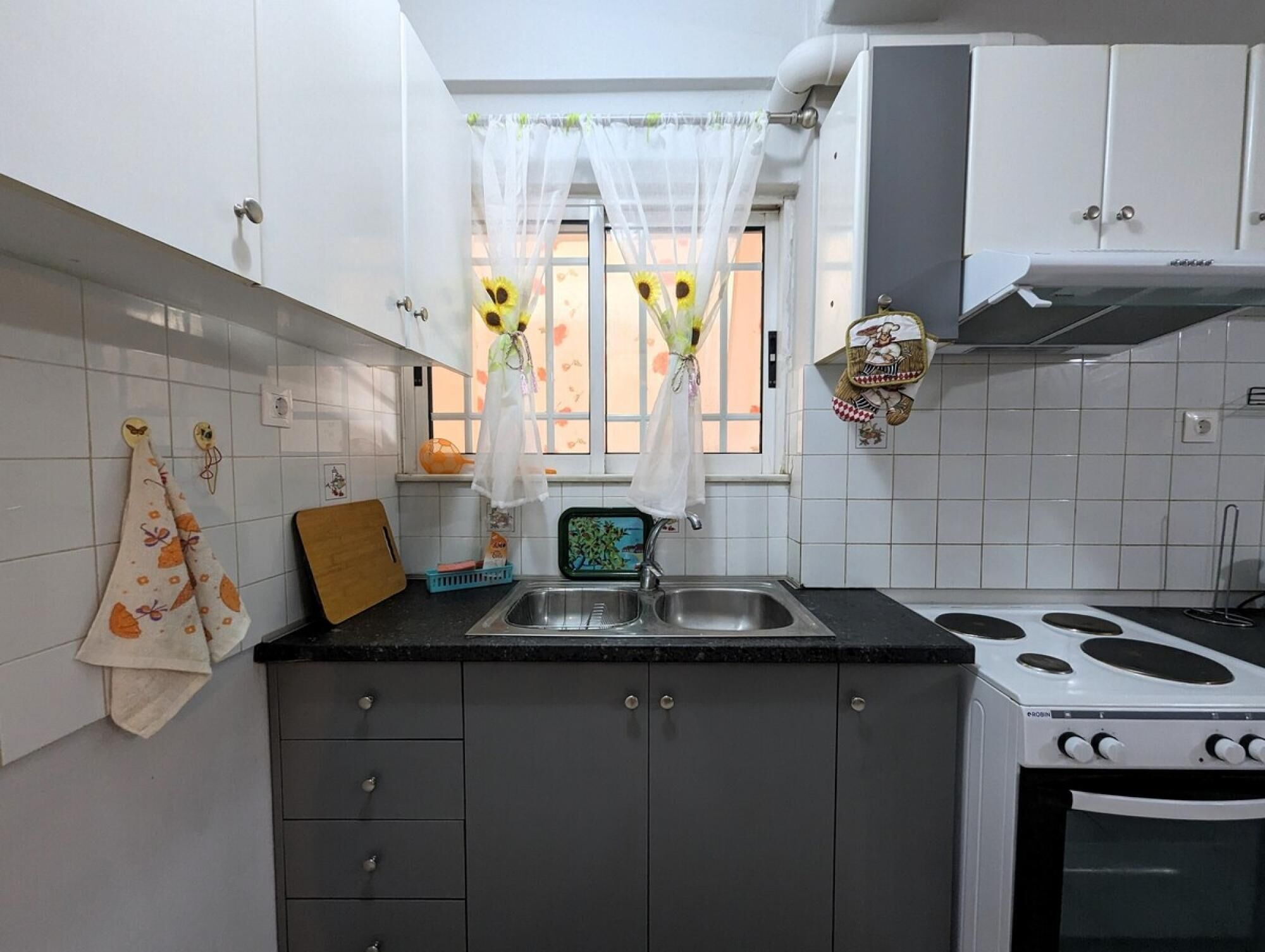 Une cuisine avec des meubles blancs, une cuisinière, un évier et une fenêtre avec un rideau.