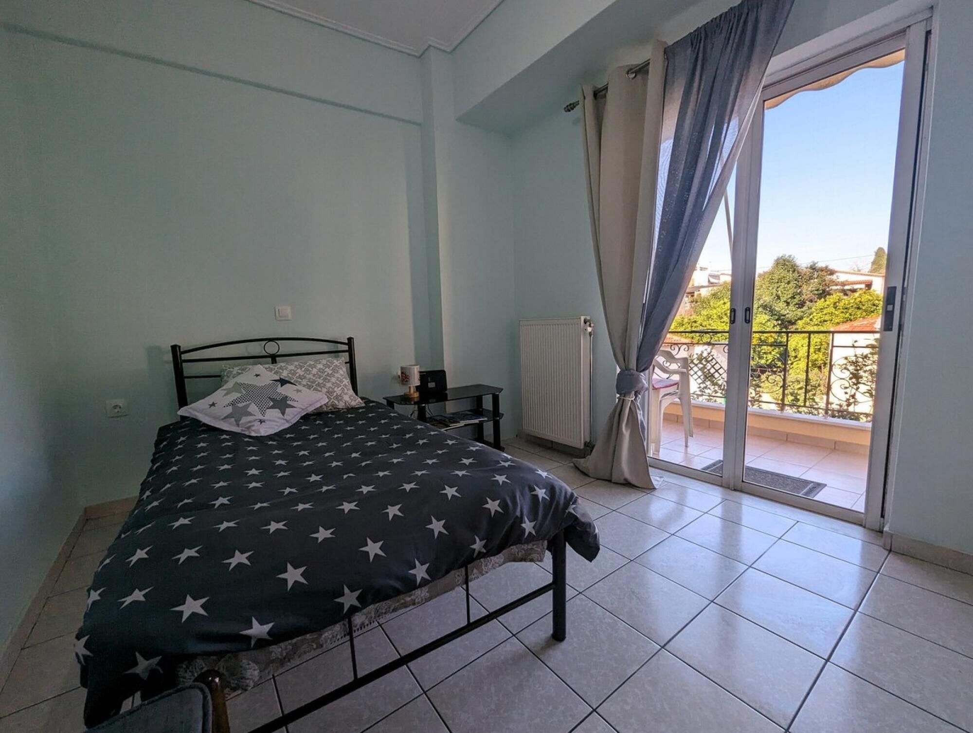 Un dormitorio con cama, mesita de noche, radiador y balcón con cortinas.