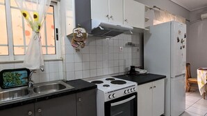 A kitchen with white cabinets, a stove, a refrigerator, and a window with a curtain. - Central Apartment in Agios Konstantinos (Kamena Vourla)