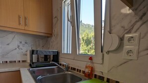 A kitchen with wooden cabinets, a sink, a green cutting board, and a window with a view of greenery outside. - Bella Vista Luxury Apartment (Kamena Vourla)