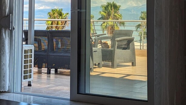 A room with large sliding glass doors leading to a balcony with outdoor furniture and a view of the sea and mountains. - Bella Vista Luxury Apartment (Kamena Vourla)