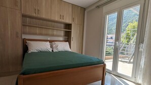 A bedroom with a wooden bed, a green bedspread, and a large window with a view of a balcony and greenery. - Bella Vista Luxury Apartment (Kamena Vourla)