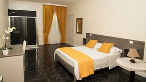 A hotel room with a bed, a desk, a chair, a small table, a mirror, and a shower area. - B&B Laura (Terzigno)