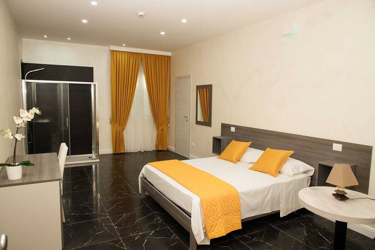 Une chambre d’hôtel comprenant un lit, un bureau, une chaise, une petite table, un miroir et un espace douche.