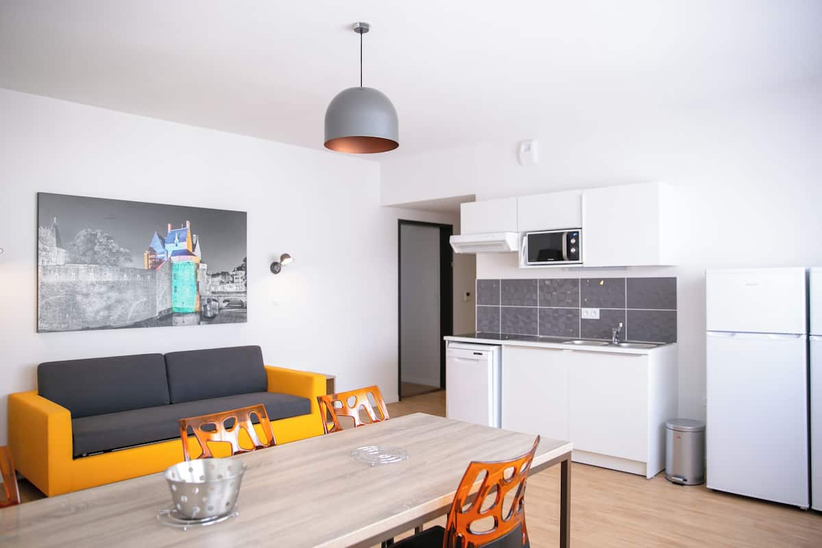Eine moderne Küche mit weißen Schränken, einer Mikrowelle und einem Kühlschrank. Es gibt einen Essbereich mit einem Holztisch und orangefarbenen Stühlen. Eine graue Pendelleuchte hängt über dem Tisch.