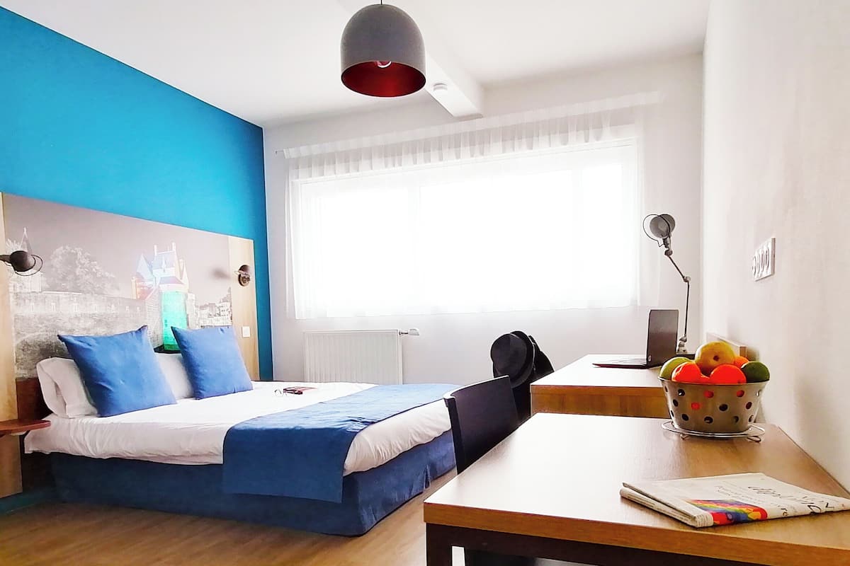 Ein Hotelzimmer mit einem Bett, einem Schreibtisch, einem Stuhl, einer Lampe, einem Fenster mit Vorhängen und einer blauen Wand.