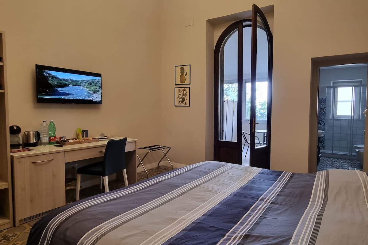 Habitación de hotel con cama, escritorio, silla, televisor y puerta que da a un balcón.