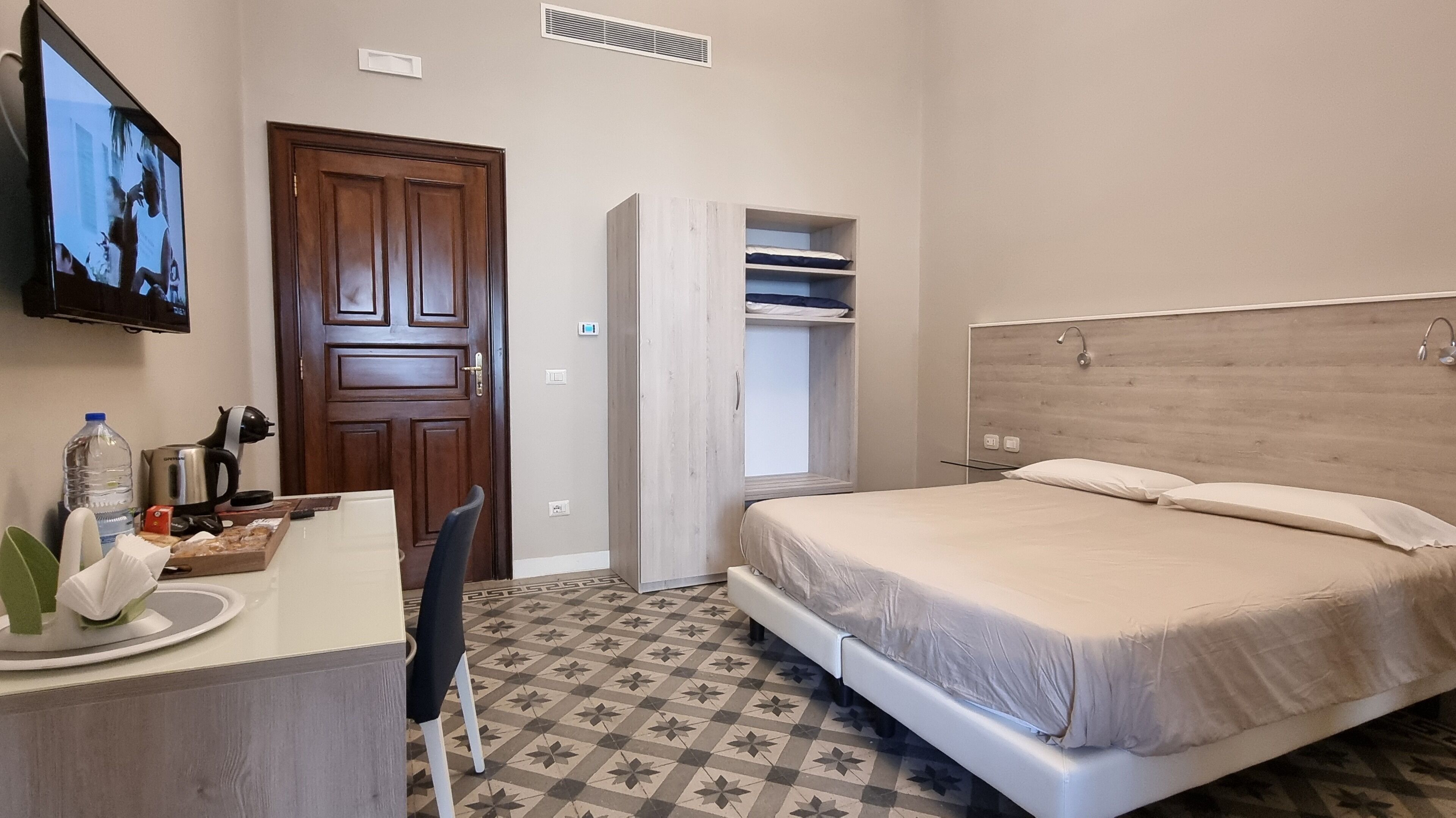 Quarto de hotel com cama, escrivaninha com cadeira, guarda-roupa e televisão na parede.