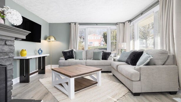 A living room with a grey sofa, a wooden coffee table, a stone fireplace, and a view of a building through large windows.