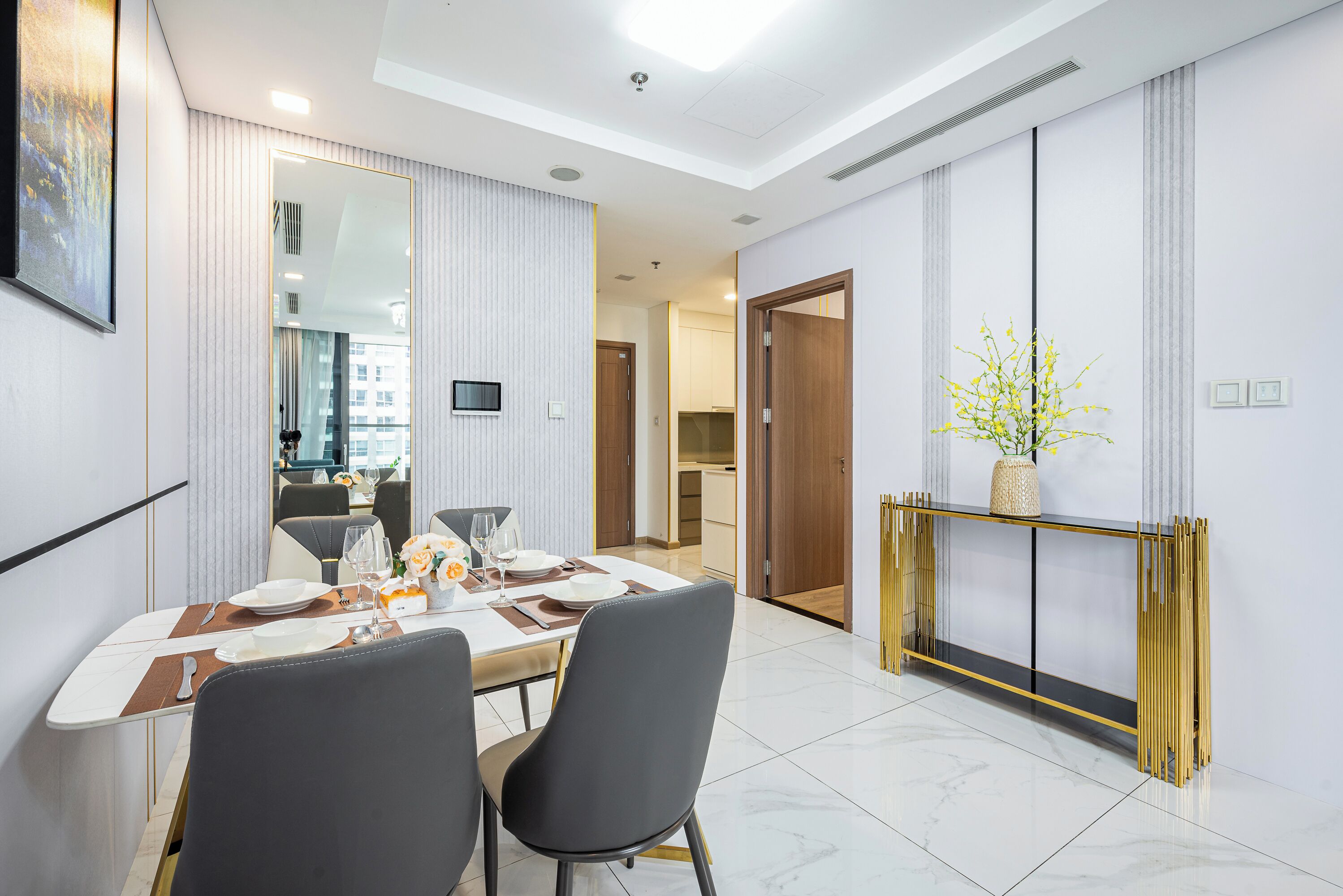 Un comedor moderno con una mesa blanca dispuesta para dos personas, un espejo grande y una consola con un florero de flores amarillas.