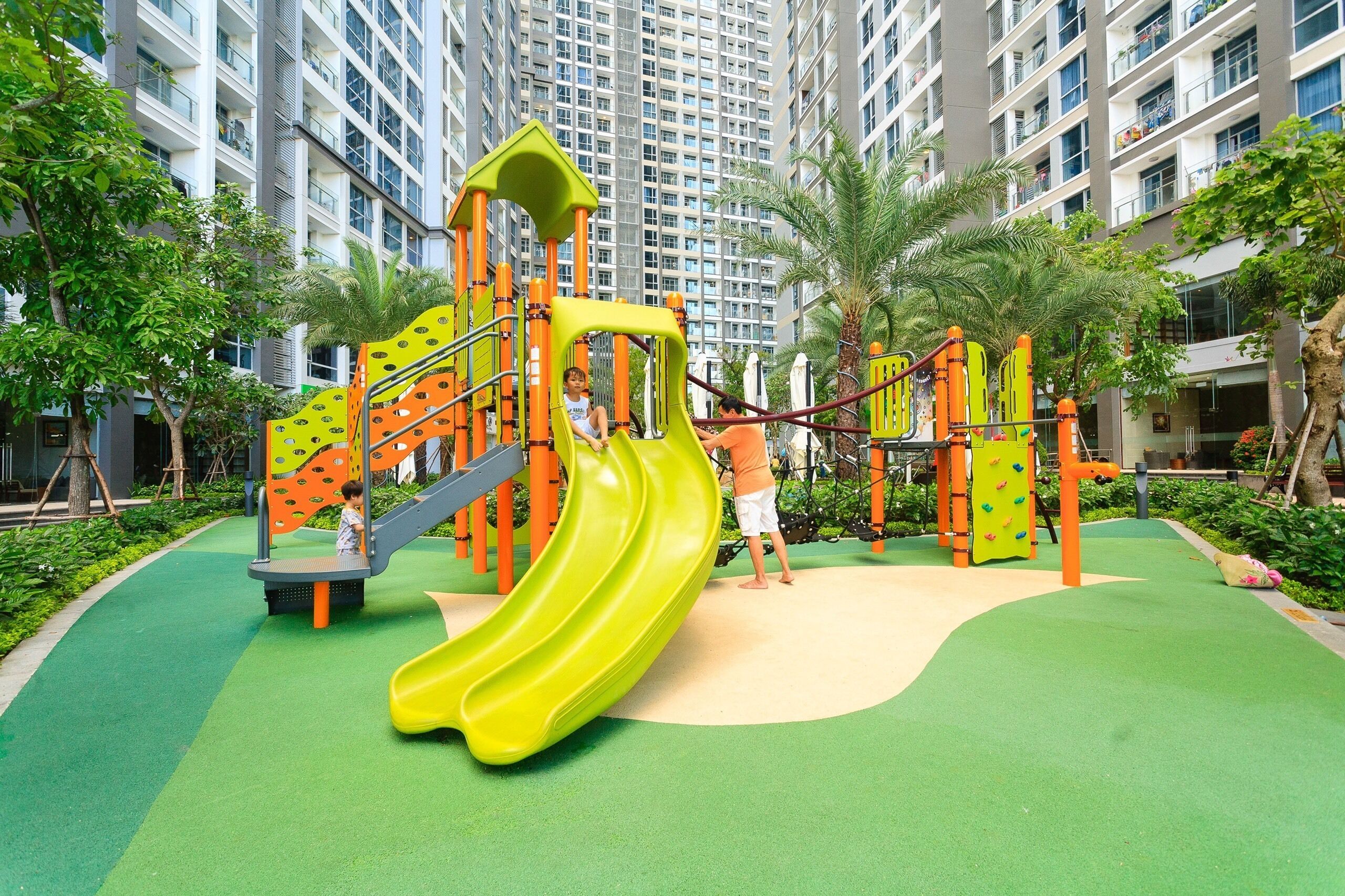 A playground with a yellow slide and climbing structures, surrounded by greenery and tall buildings.