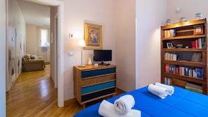 Ein Schlafzimmer mit einem Bett, einem Schreibtisch mit Computer, einem Bücherregal und einer Tür zu einem anderen Raum.