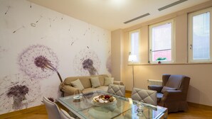 Ein modernes Wohnzimmer mit einem Glastisch, einer Couch und einem Sessel. Zwei Fenster mit Jalousien, ein Heizkörper und eine Wand mit einem Löwenzahnmuster sind zu sehen.