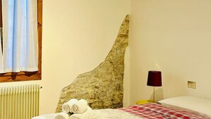 A bedroom with a stone headboard, a bed with a red and white checkered blanket, a rolled white towel, a red lamp, and a window with a white curtain. - San Pancrazio Home - Treviso (Treviso)