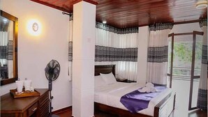 A bedroom with a bed, a bedside table, a fan, and a window with curtains. - Hotel Sara (Kandy)