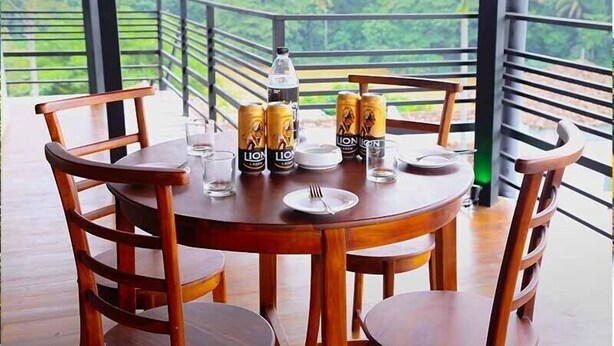 A round wooden table with four chairs, a bottle, and two cans of 'Lion' beer on a balcony with a view of greenery. - Hotel Sara (Kandy)