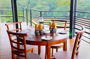 A round wooden table with four chairs, a bottle, and two cans of 'Lion' beer on a balcony with a view of greenery. - Hotel Sara (Kandy)