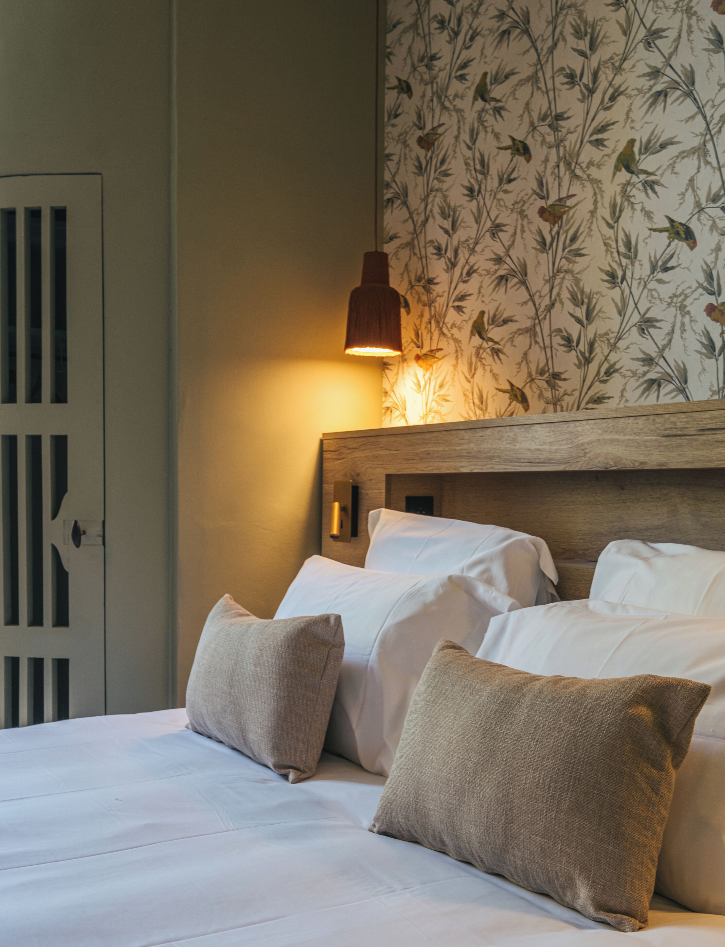 Een net opgemaakt bed met witte lakens en twee beige kussens. Een houten hoofdbord met een behangpatroon van bloemen is zichtbaar.