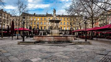 A public square with a central fountain, cobblestone pavement, and surrounding buildings with red awnings.