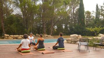 A group of people sitting on yoga mats on a wooden deck by a pool, surrounded by trees and rocks.