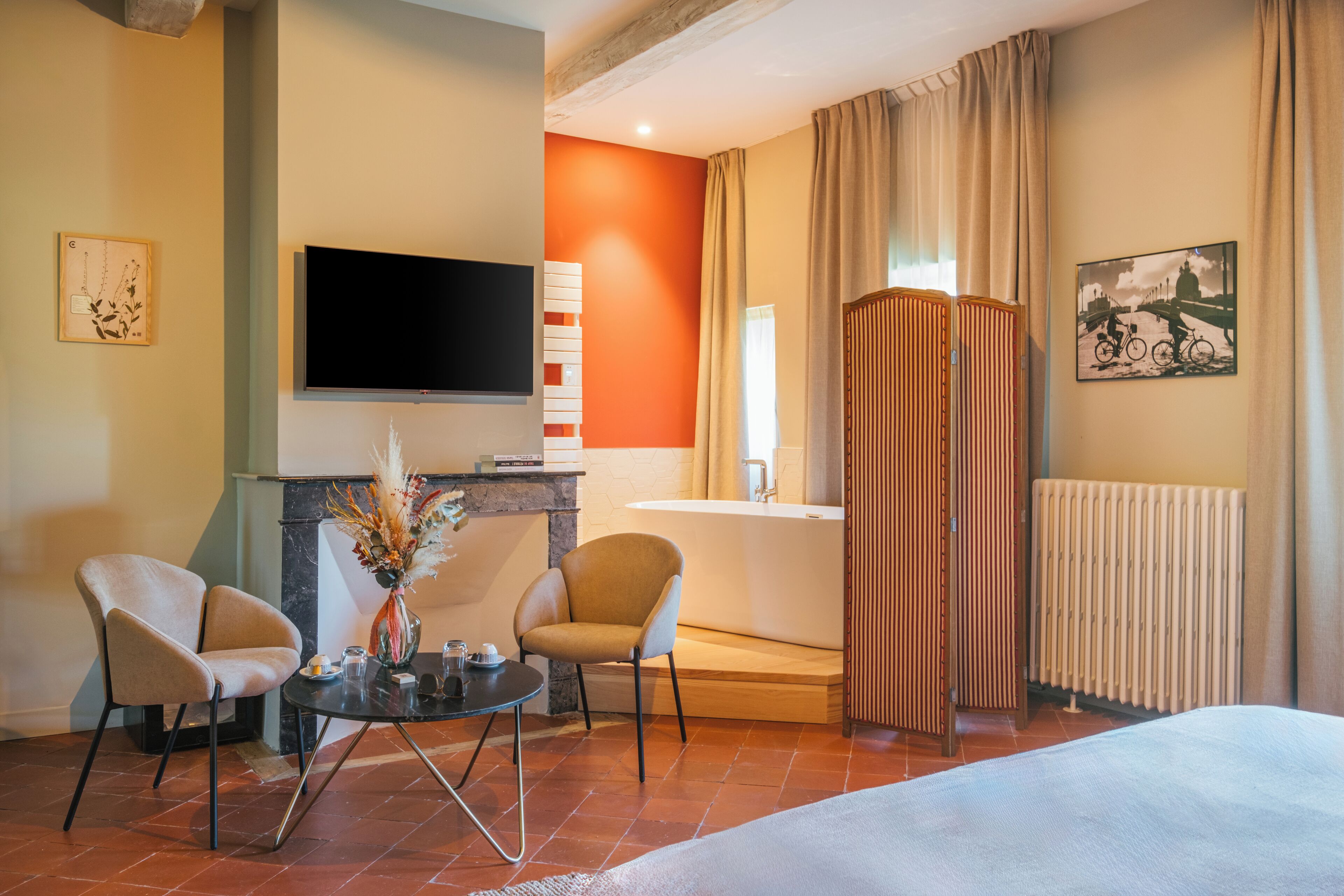 A hotel room with a flat-screen TV, a fireplace, two armchairs, a small round table, and a bed.